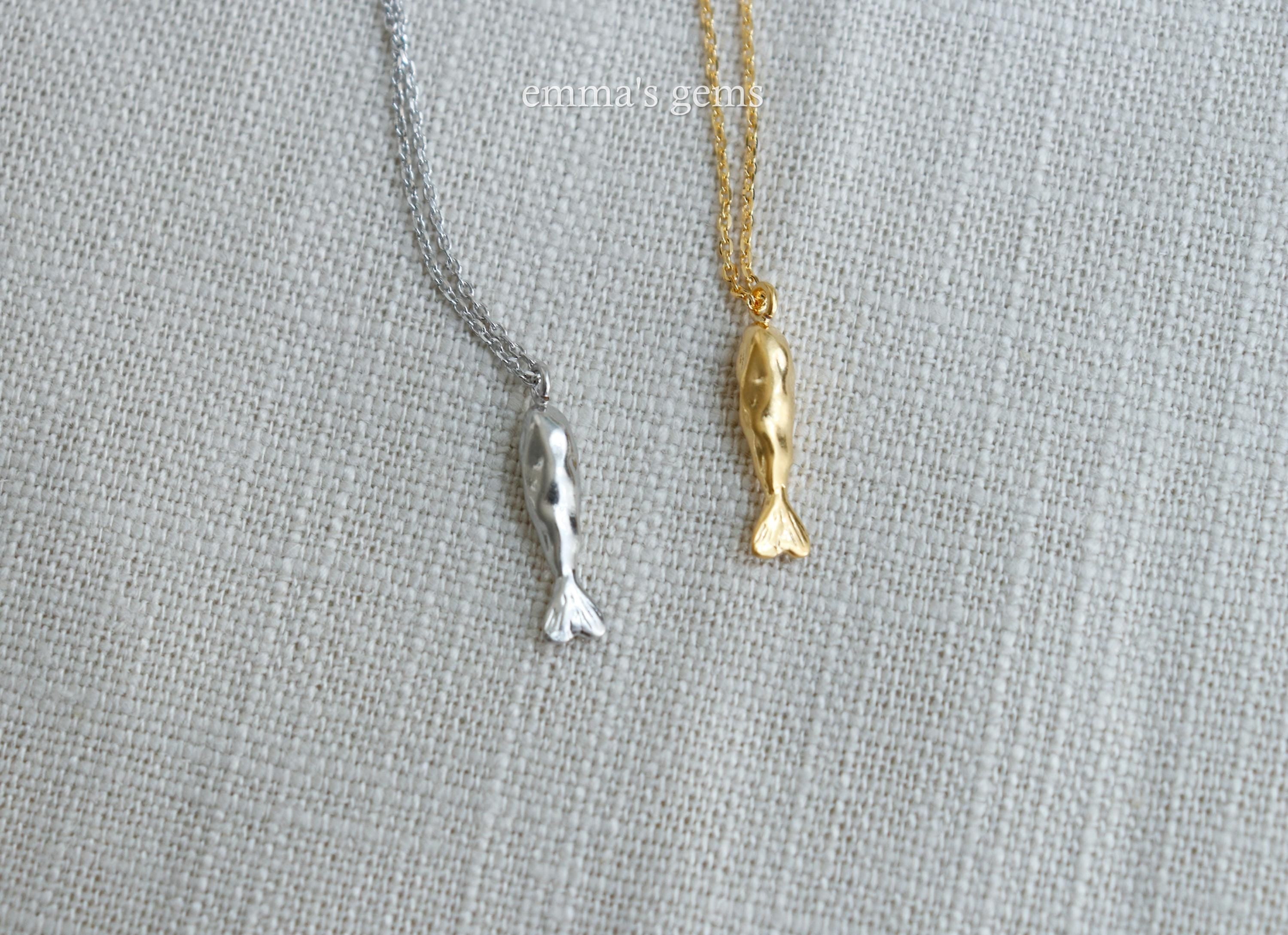 Two fish-shaped pendants, one gold and one silver, on a textured fabric background.