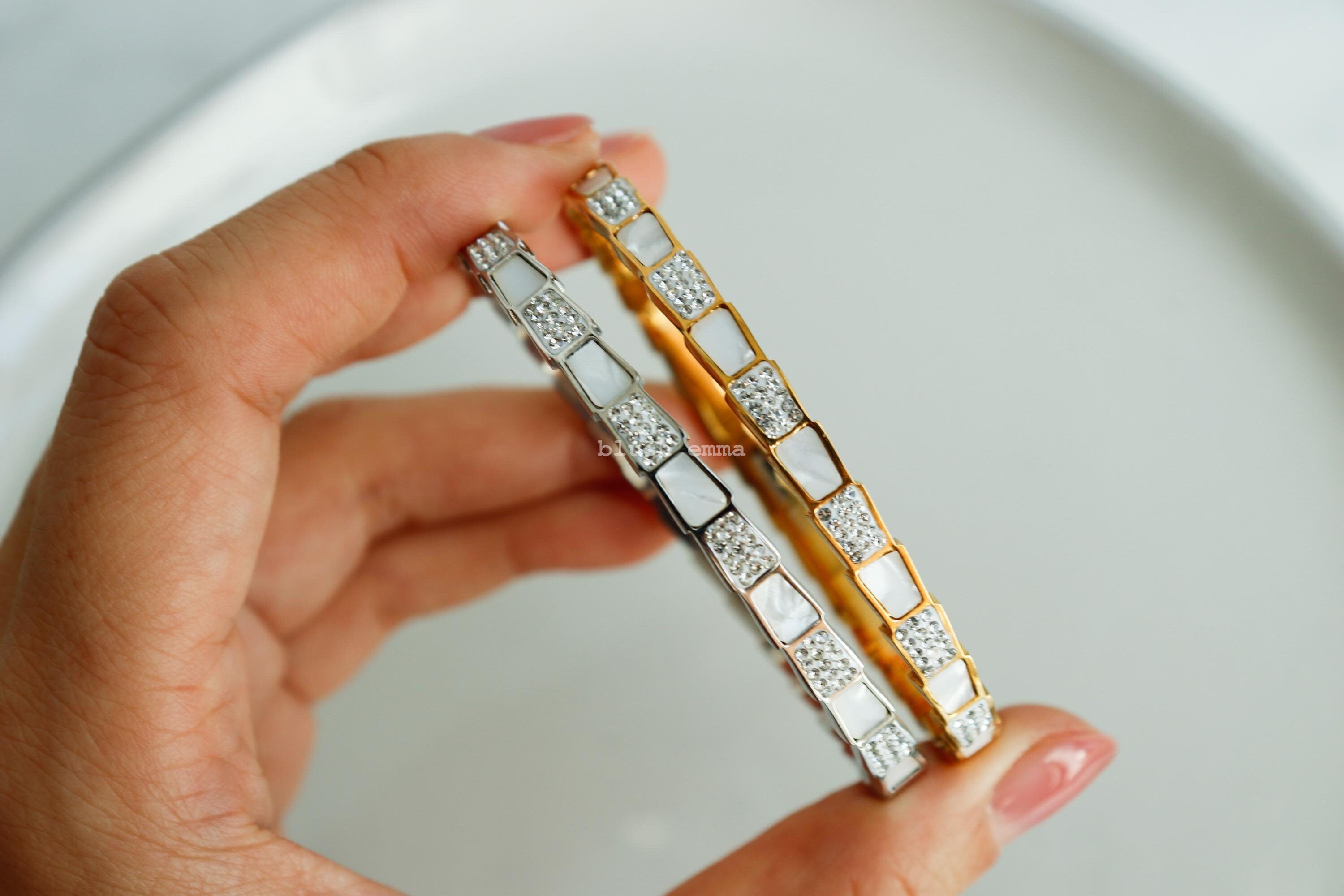 White Shell Crystal Pave Layered Trapezoid Cuff Bangle Bracelet. Stainless Steel A hand holding two layered trapezoid bangle bracelets with white shell and rhinestone pave, one with a gold finish and the other with a silver finish.