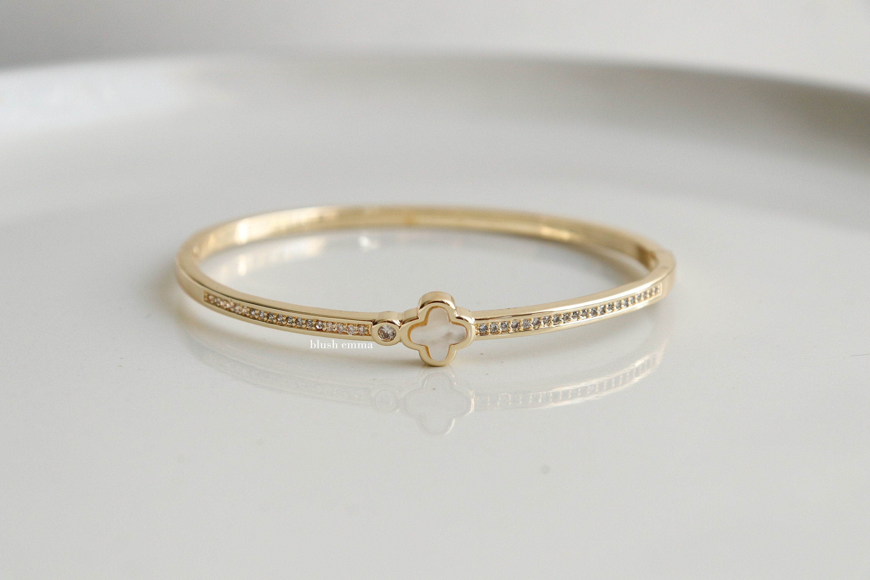 A gold-colored bangle bracelet with a mother of pearl clover at the center, studded with rhinestones on the front, displayed on a white surface.