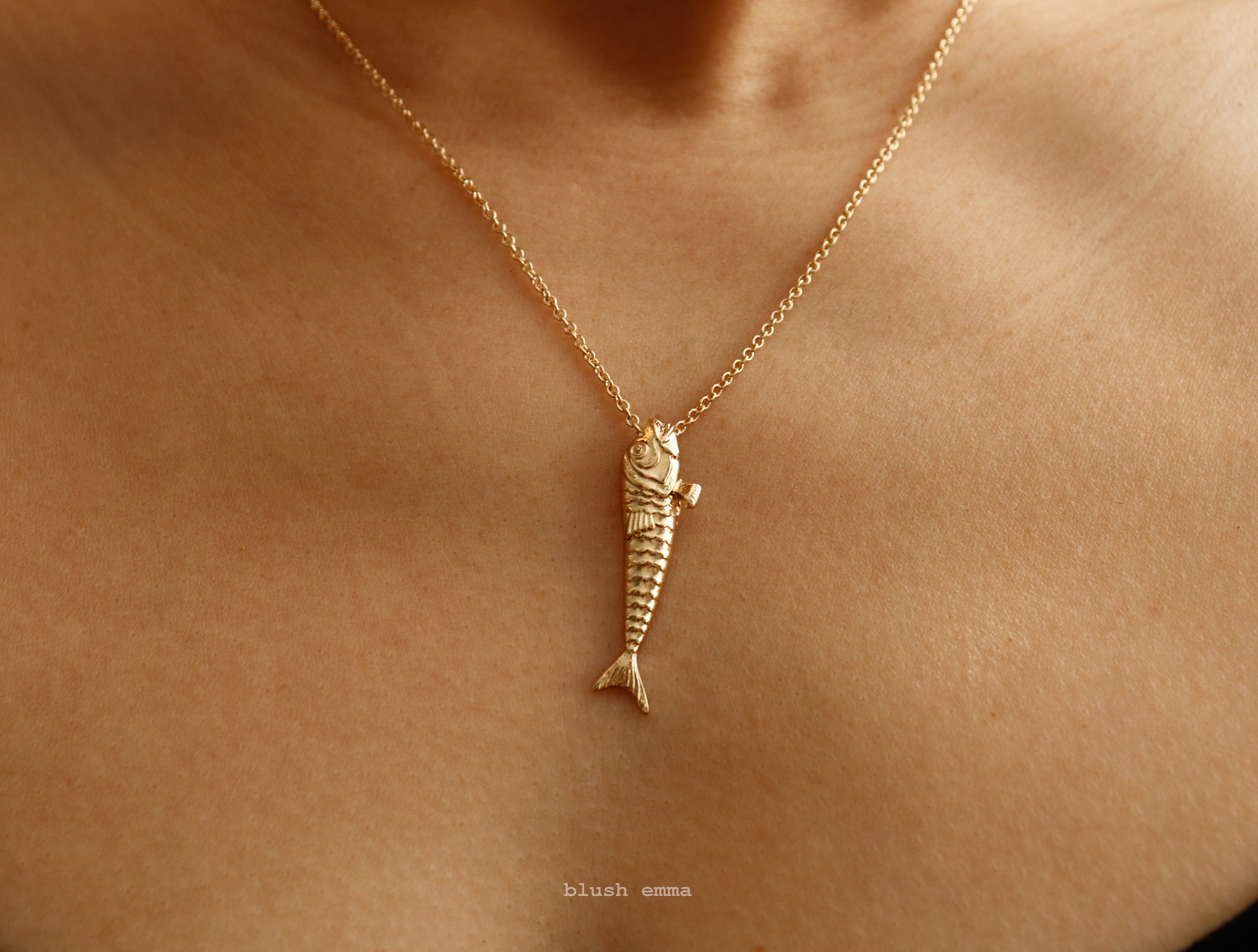 Gold fish-shaped pendant necklace on a person's neck with 'blush emma' branding.