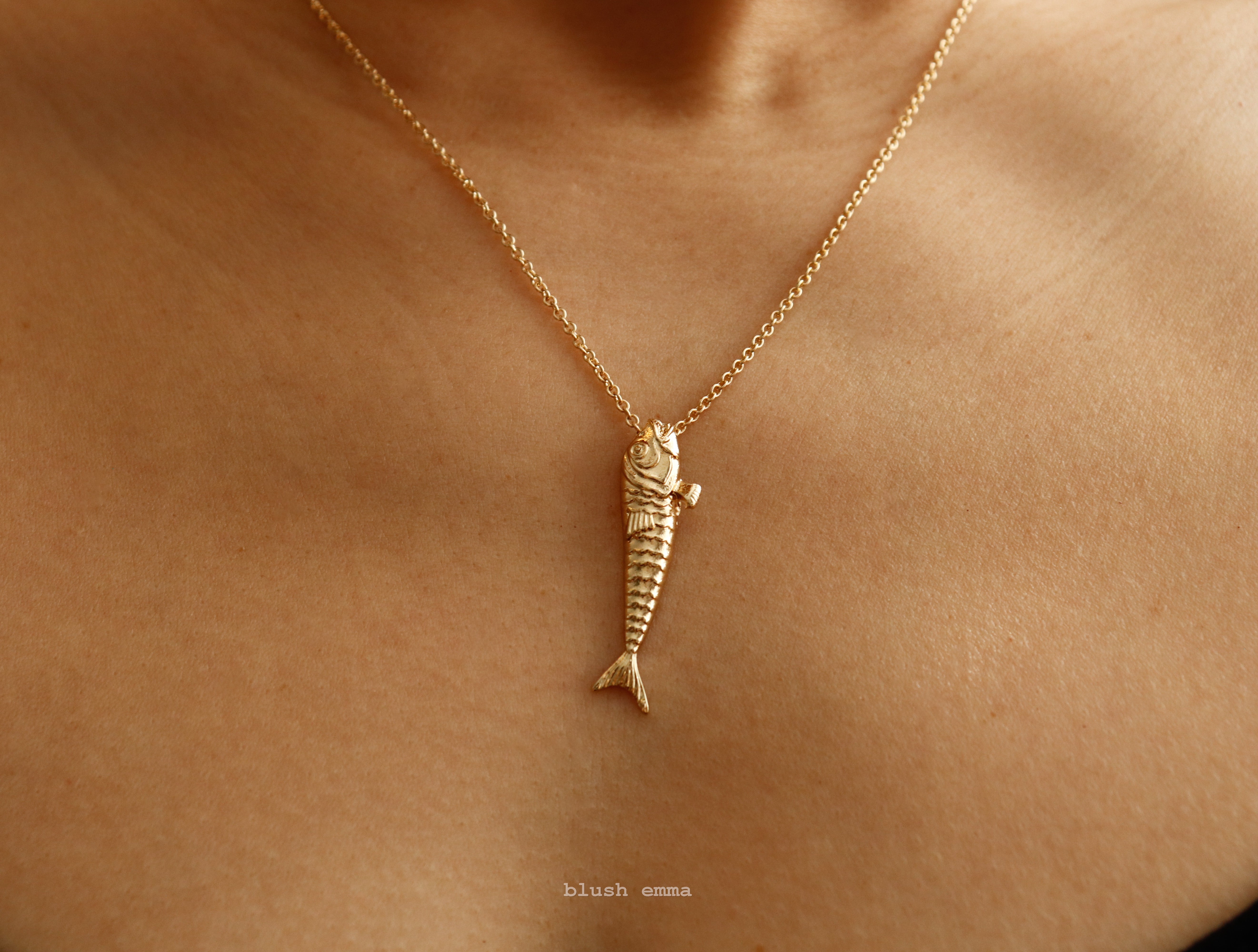 Gold fish-shaped pendant necklace on a person's neck with 'blush emma' branding.
