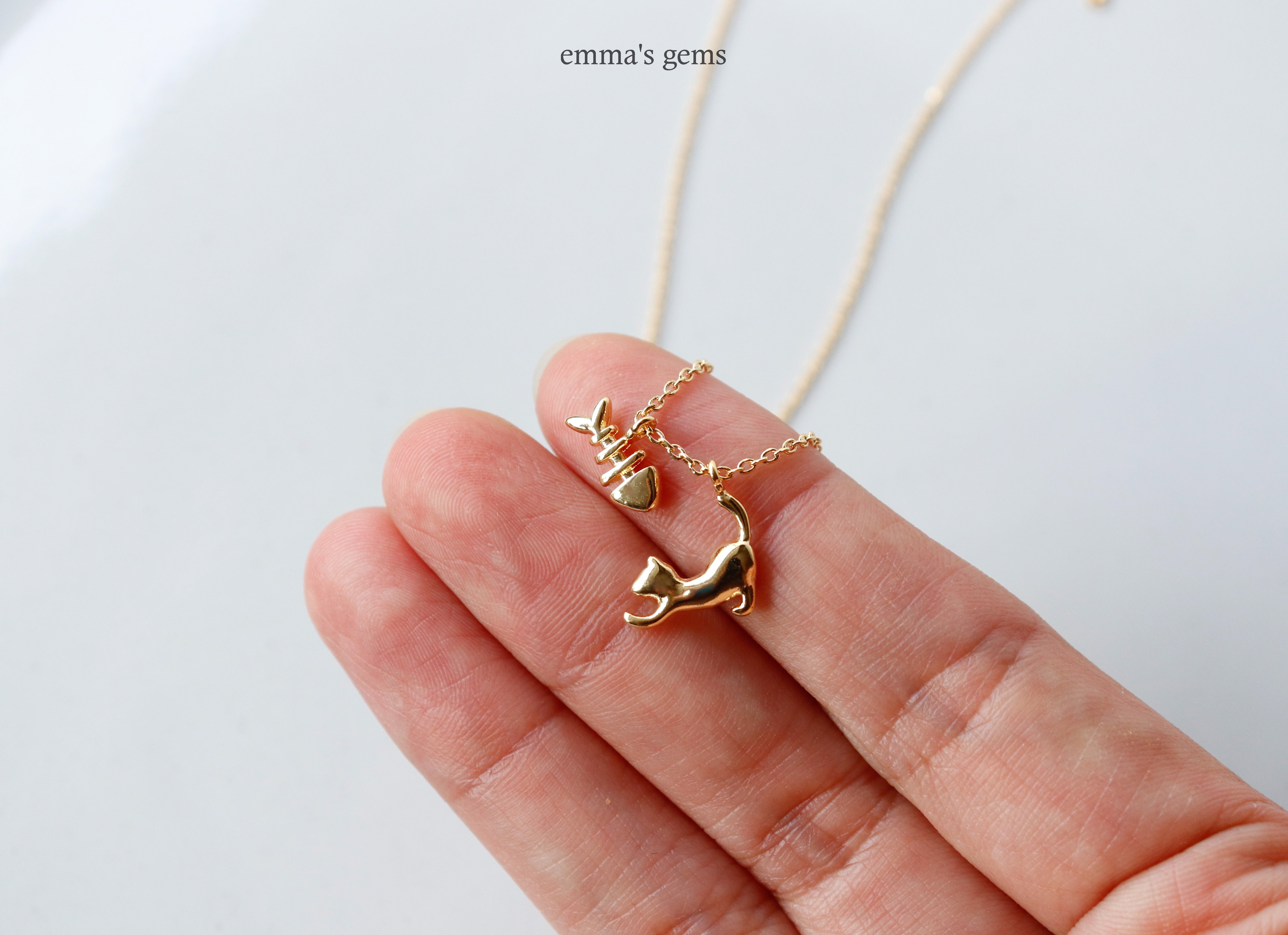 Gold cat shaped and fishbone pendant on a chain held between fingers against a white background.