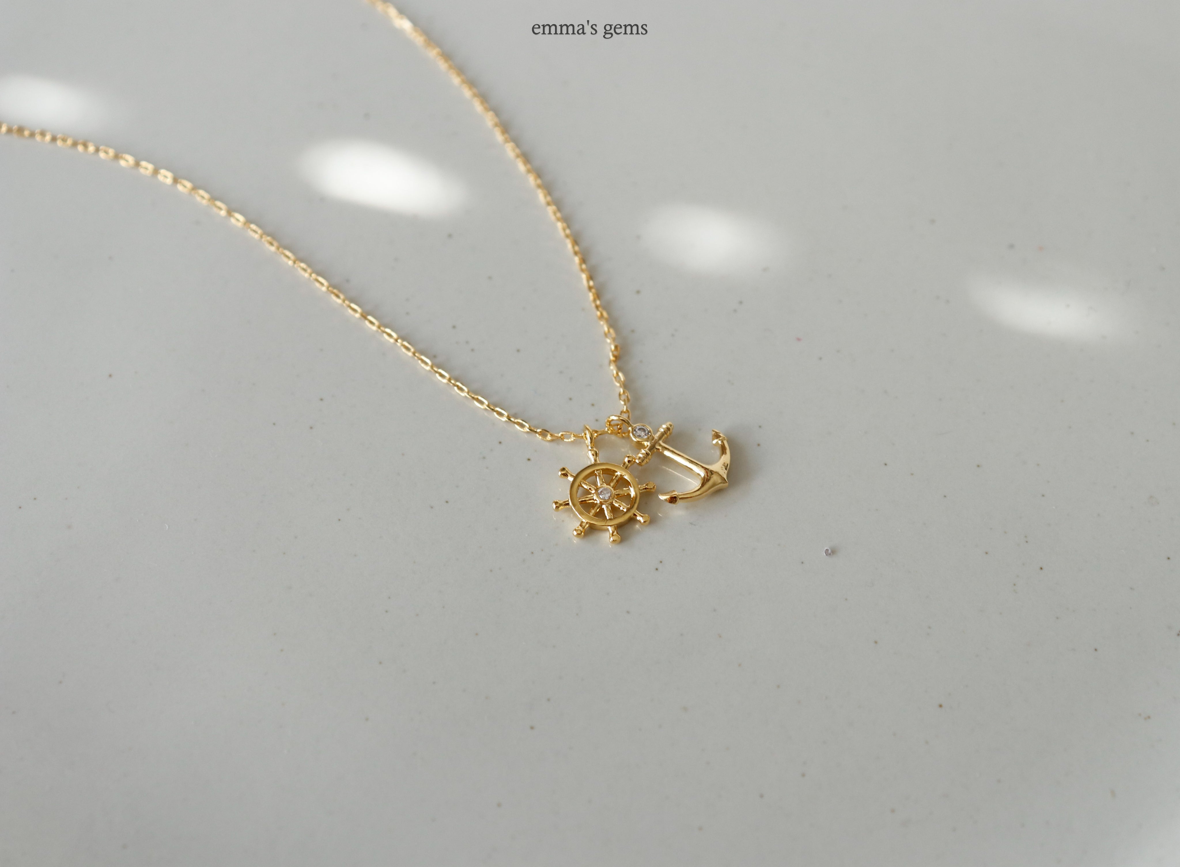 Gold necklace with a boatwheel and anchor pendant on a light gray background