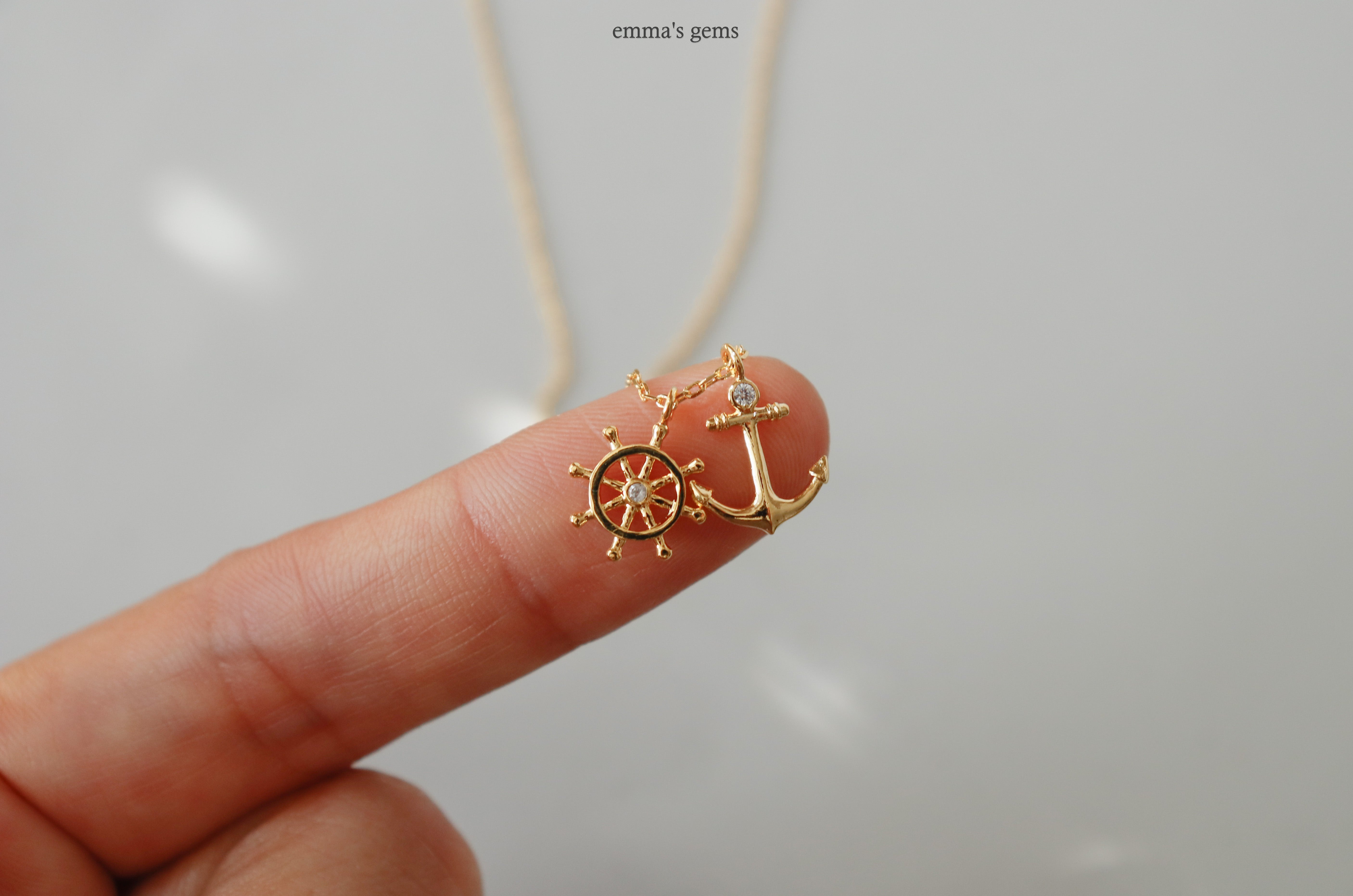 Gold anchor and wheel pendant on a necklace held by a finger against a light background.