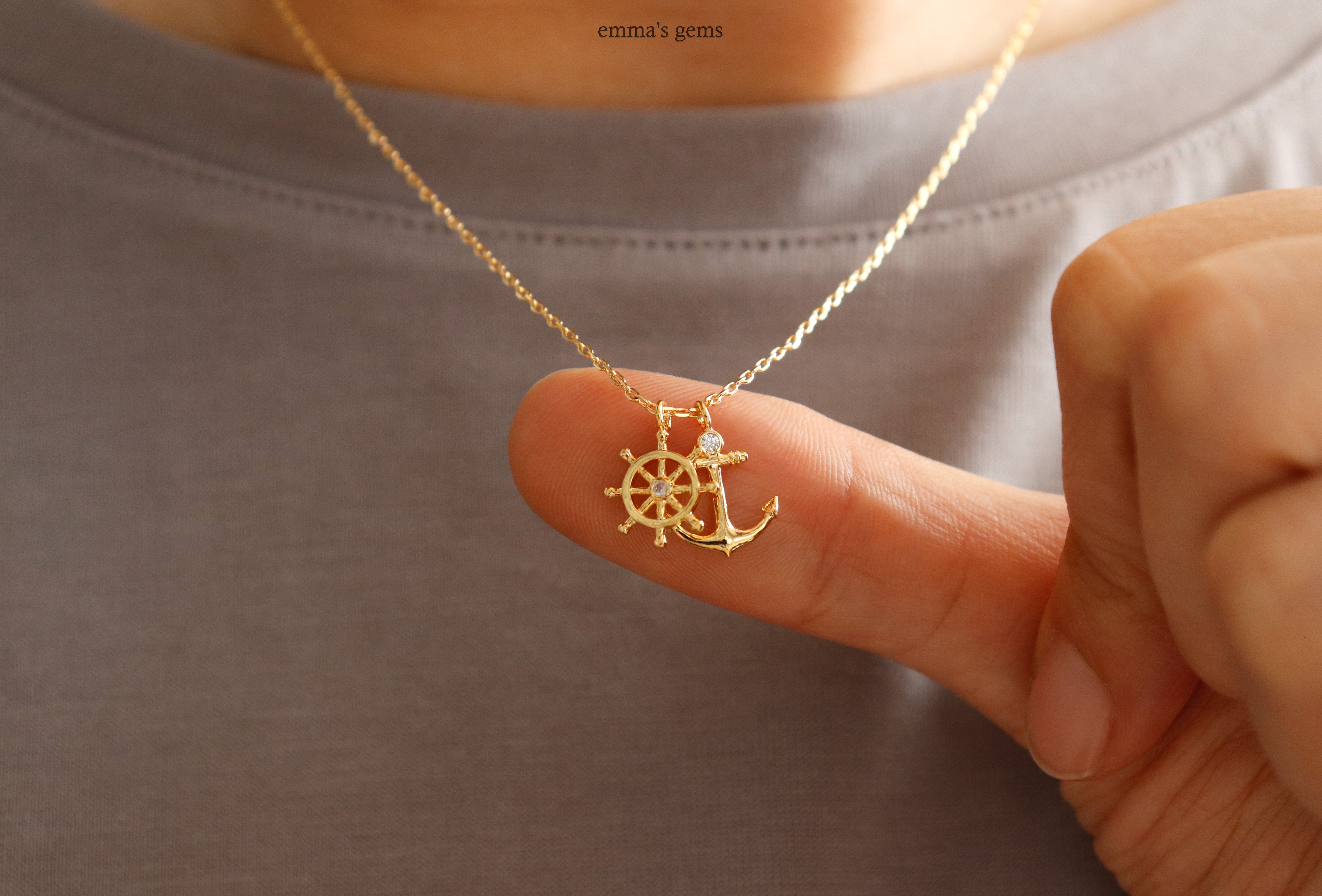 Gold necklace with a ship wheel and anchor pendant held by a hand against a neutral background
