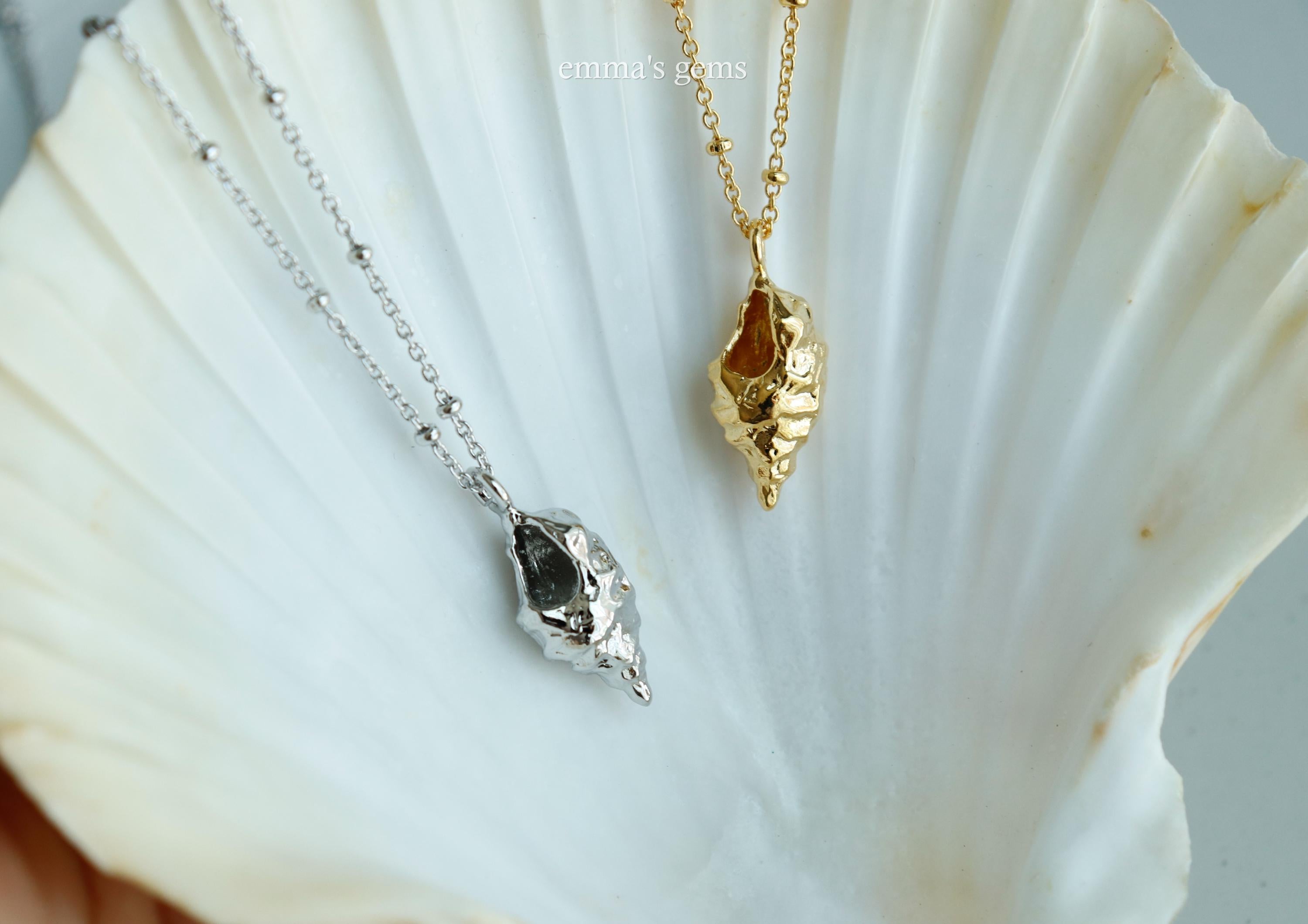 Two shell-shaped pendants, one gold and one silver, on a white shell background.