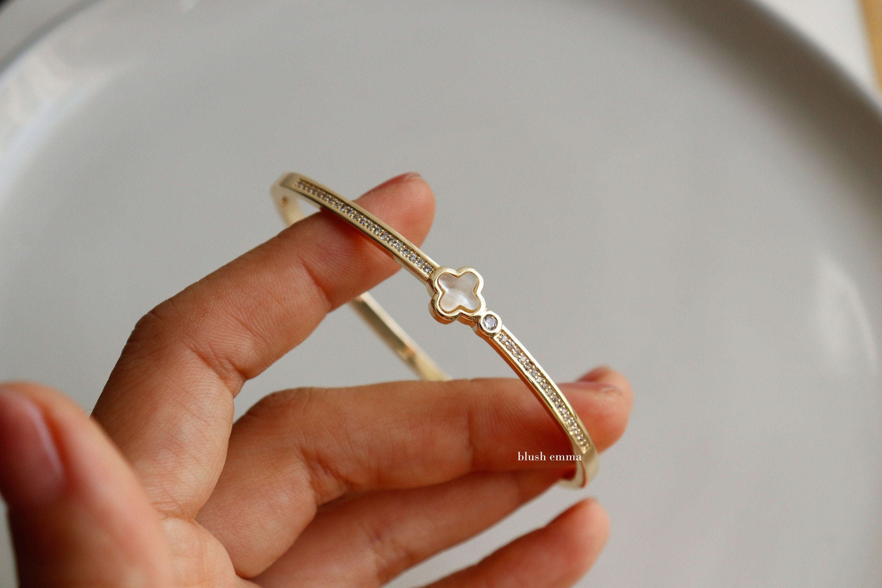 Blush Emma Mother of Pearl Clover Rhinestone Studded Bangle Bracelet A gold-colored mother of pearl clover rhinestone studded bangle bracelet with a figure 8 clamp closure, displayed on a person's hand.