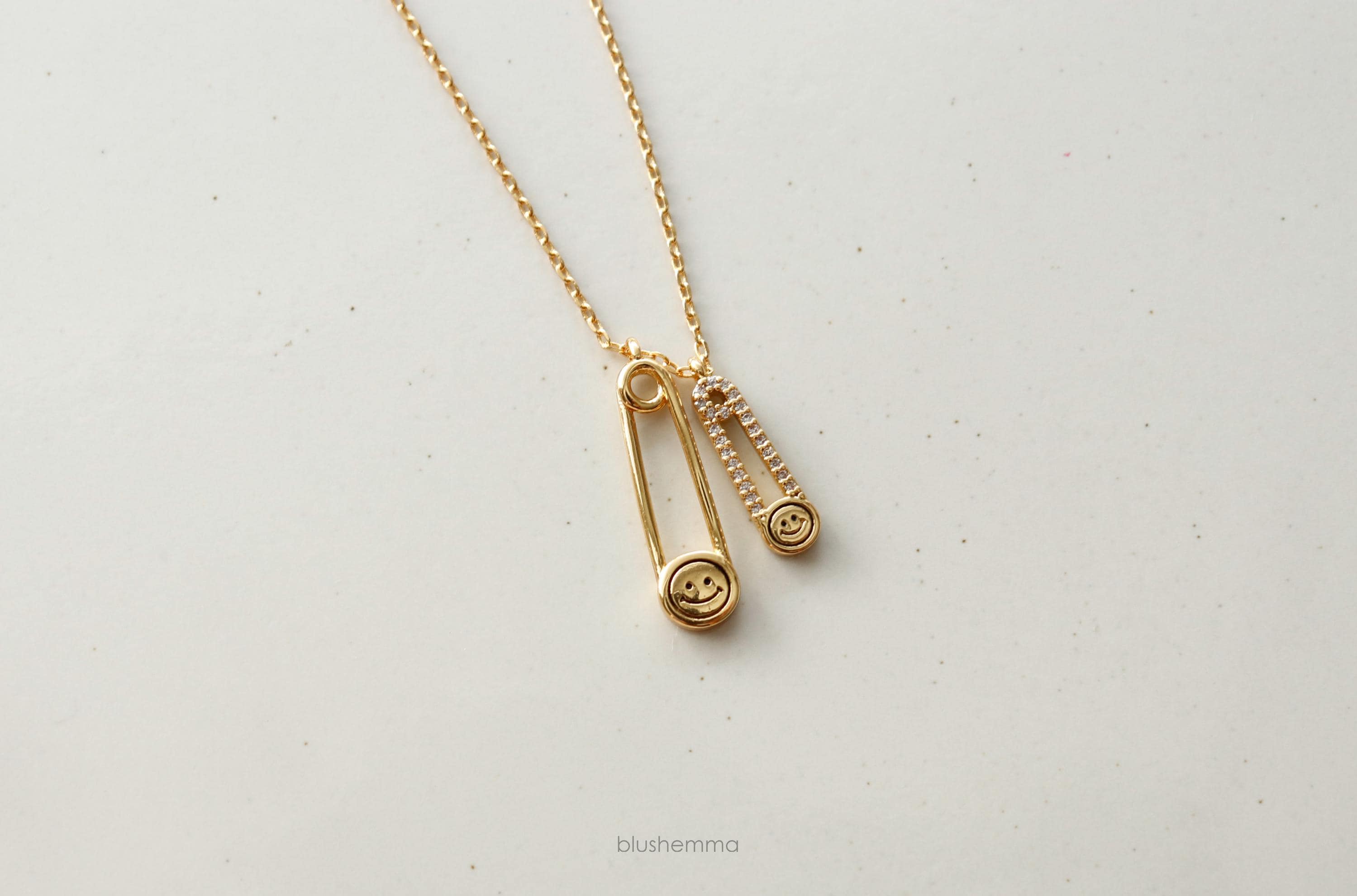Double Safety Pin Charm Necklace.
Each pin has a Happy Face and the smaller one is cubic paved along pin shape.